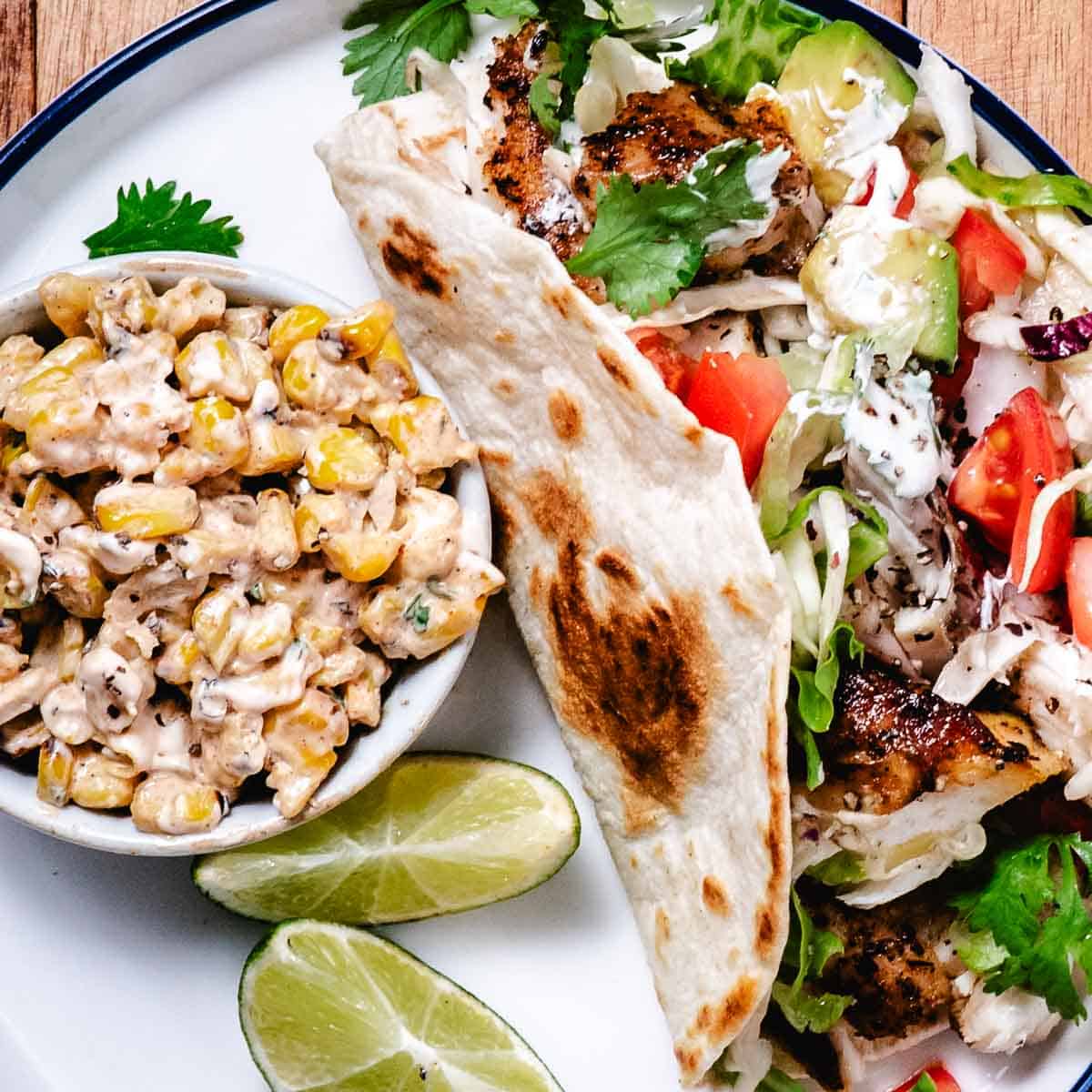 Close-up of grilled red snapper fish tacos topped with avocado, tomato, cabbage, cilantro, and crema, paired with creamy street corn salad and fresh lime wedges.