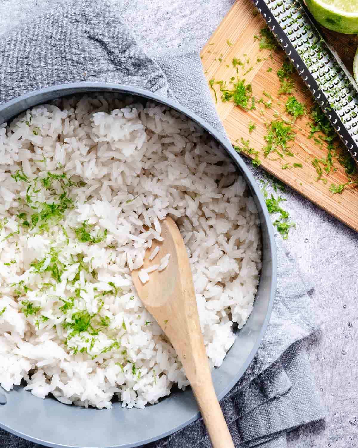 Coconut rice in a saucepan with fresh lime zest, a wooden spoon, and a lime on a cutting board.