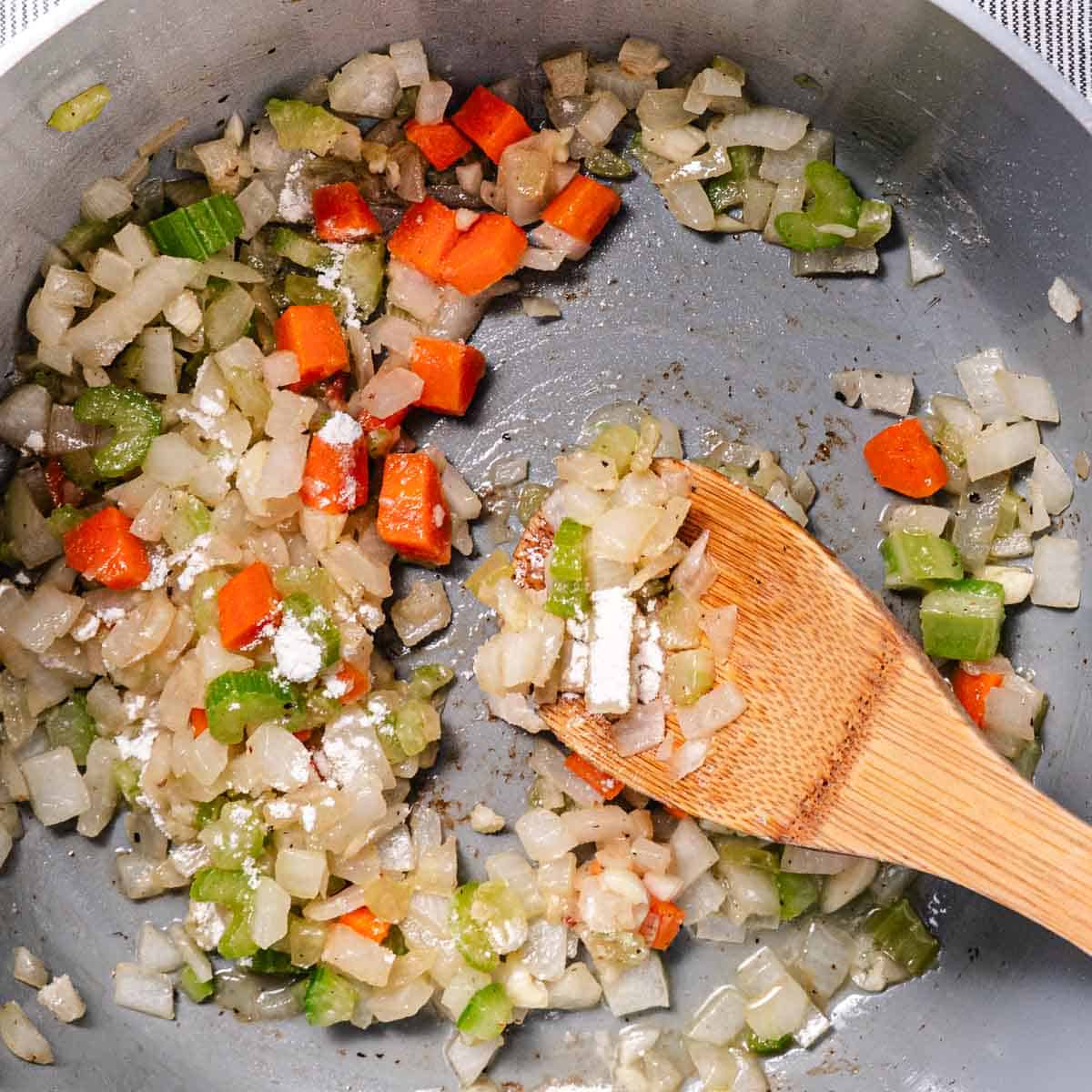 Flour being stirred into cooked onions, celery, carrots, and garlic to make a roux for creamy corn chowder.