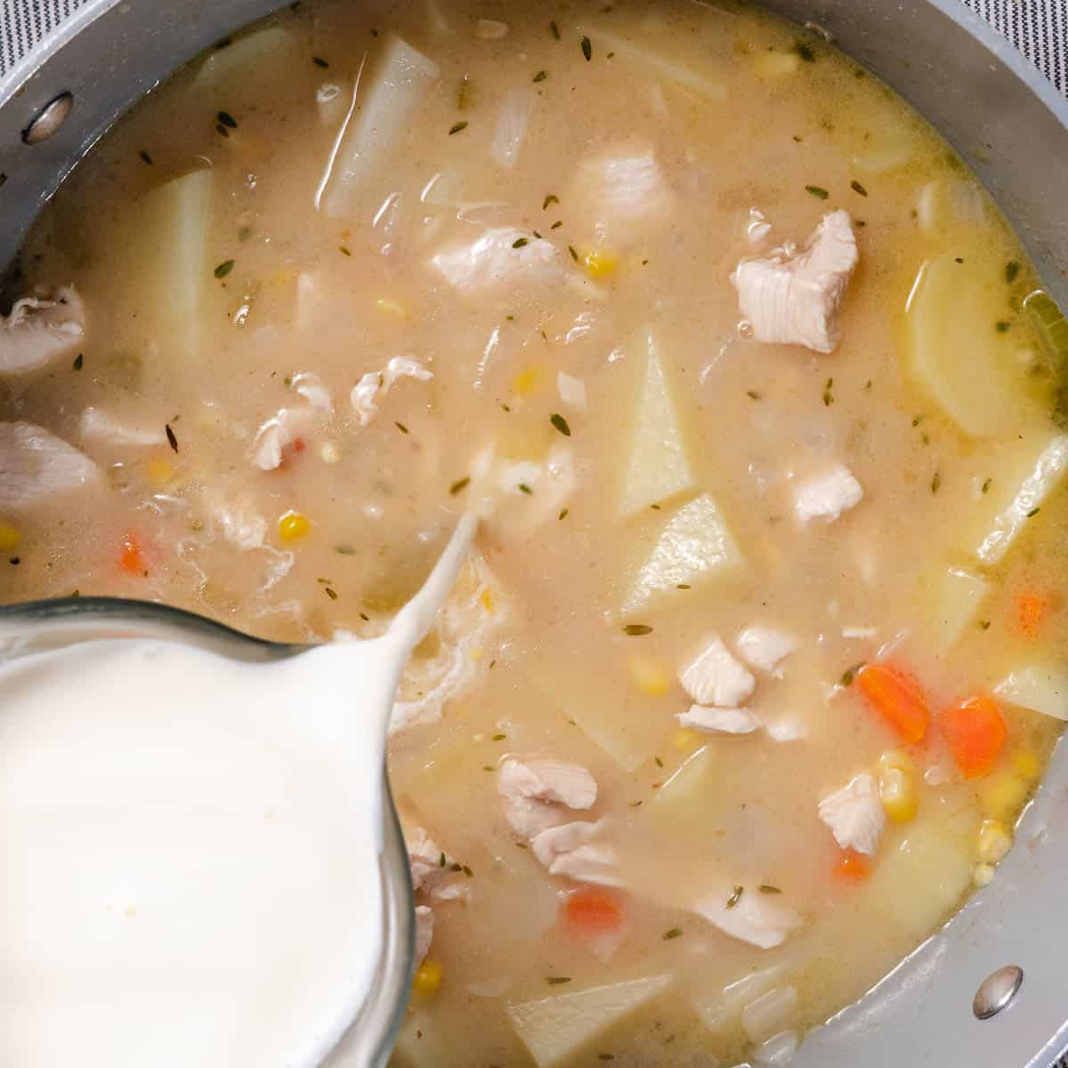 Heavy cream being poured into a pot of chicken chowder with potatoes, corn, carrots, celery, thyme, and cooked chicken.
