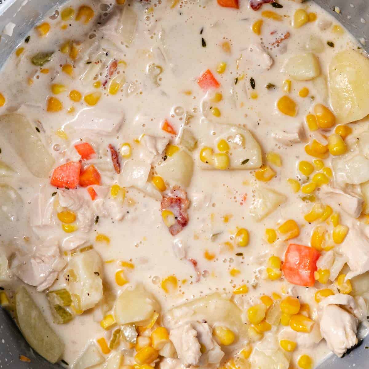 Creamy chicken chowder simmering in a pot with chicken breast, corn, potatoes, carrots, celery, bacon, and thyme.