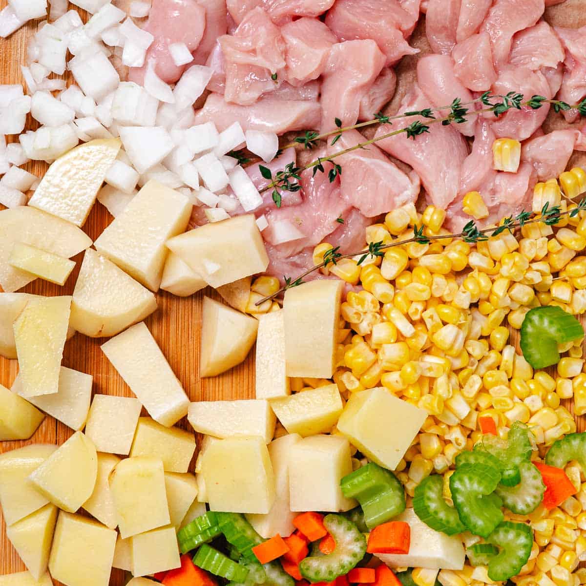Raw chicken breast, diced potatoes, corn kernels, onion, celery, carrots, and fresh thyme arranged on a cutting board for homemade chowder.