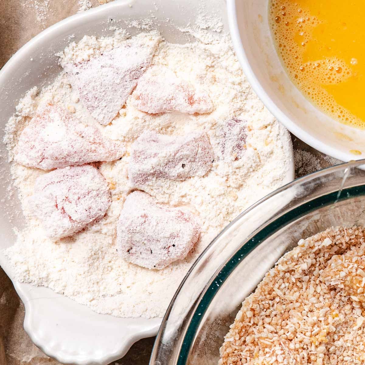 Gluten-free coconut chicken nuggets dredged in coconut flour with beaten eggs and coconut breadcrumb coating nearby.