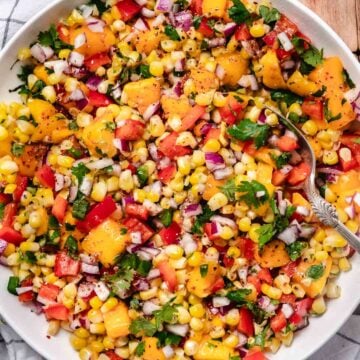 Corn mango salsa mixed in a glass bowl with diced mango, sweet corn, red onion, red bell pepper, jalape&ntilde;o, cilantro, lime zest, and a spoon.