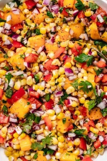 Corn mango salsa mixed in a glass bowl with diced mango, sweet corn, red onion, red bell pepper, jalape&ntilde;o, cilantro, lime zest, and a spoon.