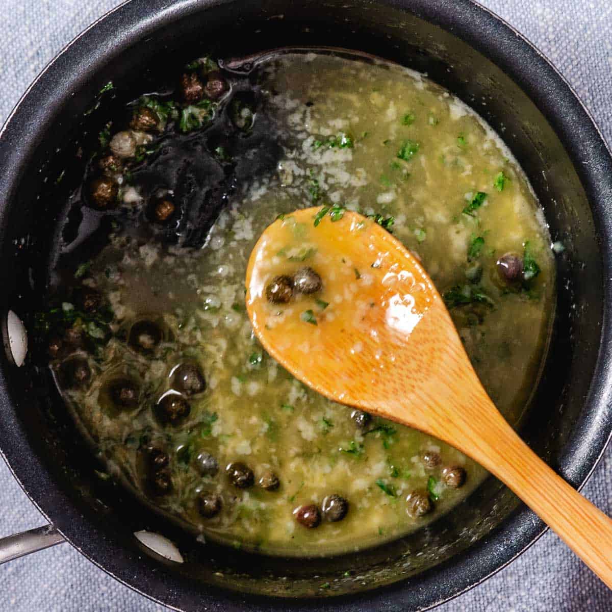 Lemon butter caper sauce with garlic and parsley in a skillet, stirred with a wooden spoon.