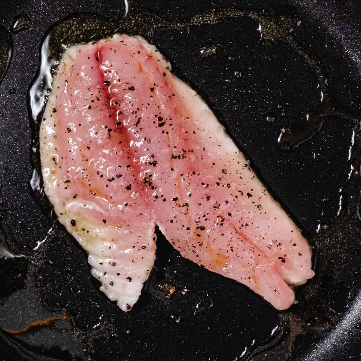 Seasoned white fish fillet cooking in oil in a black skillet.