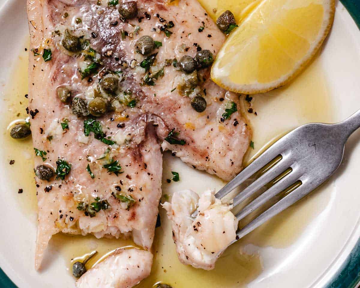 Flaky seared fish with lemon butter caper sauce, with a fork lifting a bite from the plate.