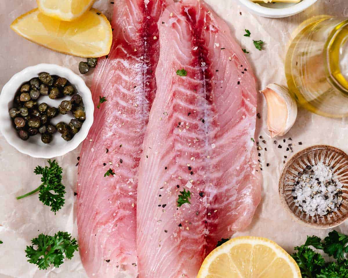 Raw fish fillets with lemon, capers, garlic, parsley, olive oil, and coarse salt arranged on parchment paper.