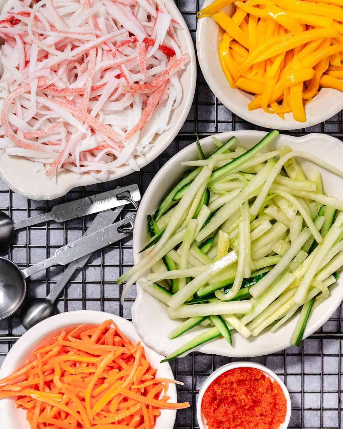 Shredded imitation crab, sliced mango, cucumber, carrots, and masago arranged in small white dishes for mango kani salad.