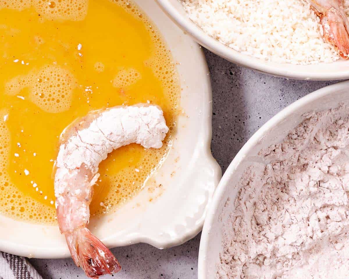 Raw shrimp dipped in egg wash beside bowls of cornstarch and coconut flakes for gluten-free coconut shrimp.