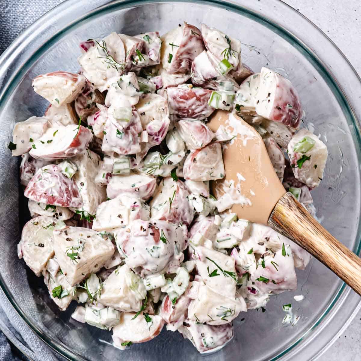 Red potato salad with celery, red onion, fresh dill, and creamy dressing being mixed in a glass bowl.