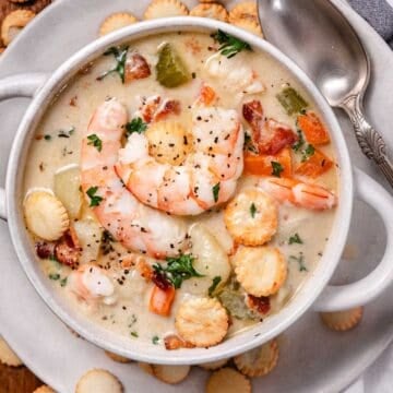 Creamy shrimp chowder with bacon, potatoes, carrots, celery, parsley, and oyster crackers in a white bowl.