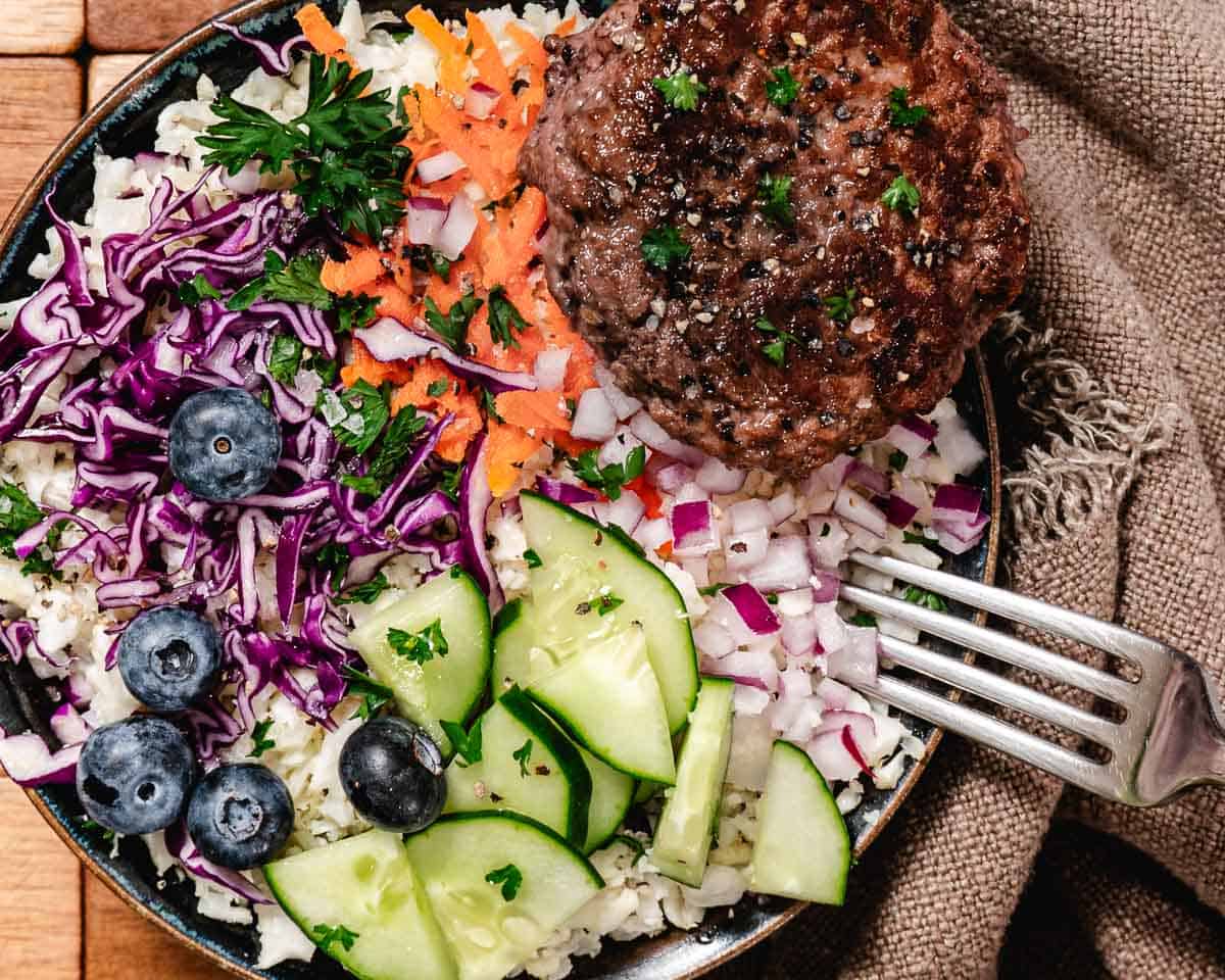 burger bowl and fork with cauliflower rice, cabbage, onions, carrots, cucumbers, and blueberries