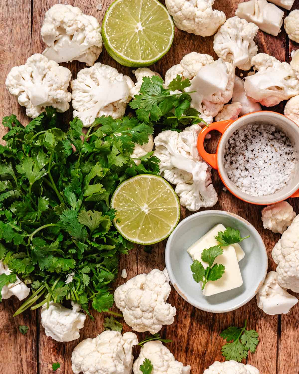 Fresh cauliflower florets, cilantro, lime halves, butter, kosher salt, and black pepper arranged on a wooden board for making cilantro lime cauliflower rice.