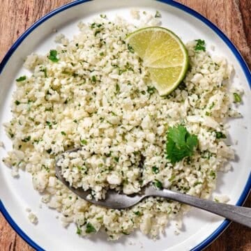 Cilantro lime cauliflower rice served on a white plate with a lime wedge, fresh cilantro, and a spoonful ready to serve.
