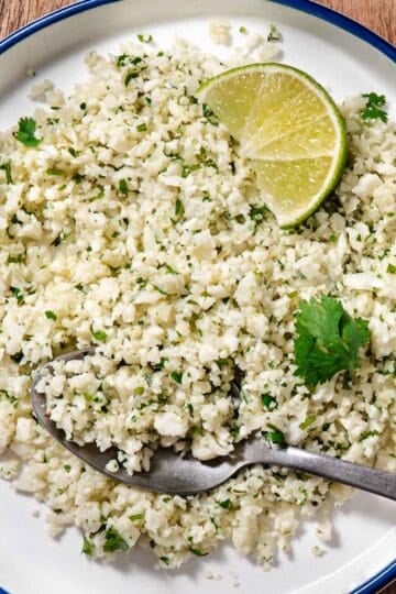 Cilantro lime cauliflower rice served on a white plate with a lime wedge, fresh cilantro, and a spoonful ready to serve.