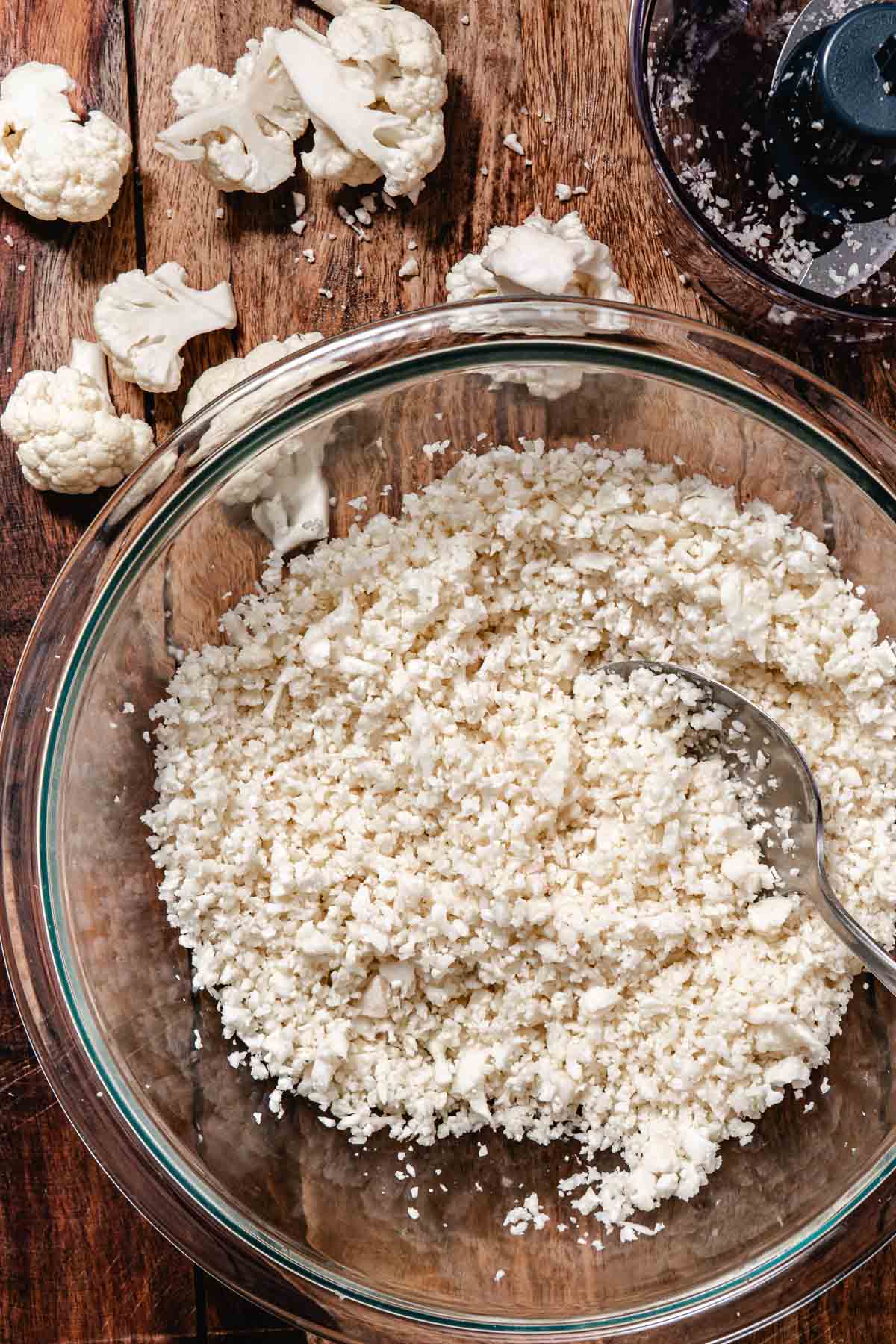 Fresh cauliflower rice in a glass bowl with cauliflower florets and a food processor nearby.