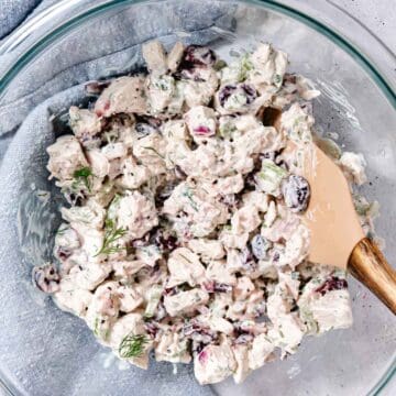 Turkey salad with dried cranberries, celery, red onion, and fresh herbs being mixed in a glass bowl.