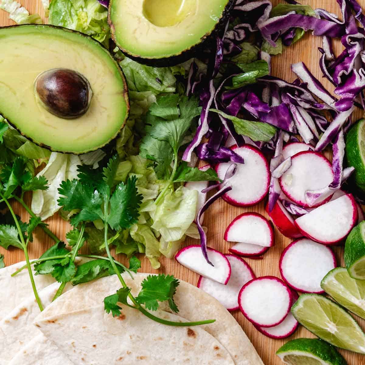Fish taco toppings on a wooden board with avocado, shredded cabbage, lettuce, radishes, cilantro, lime wedges, and flour tortillas.