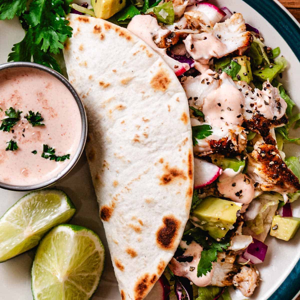 Blackened fish taco salad served with a warm flour tortilla, avocado, radishes, lime wedges, and creamy taco sauce.