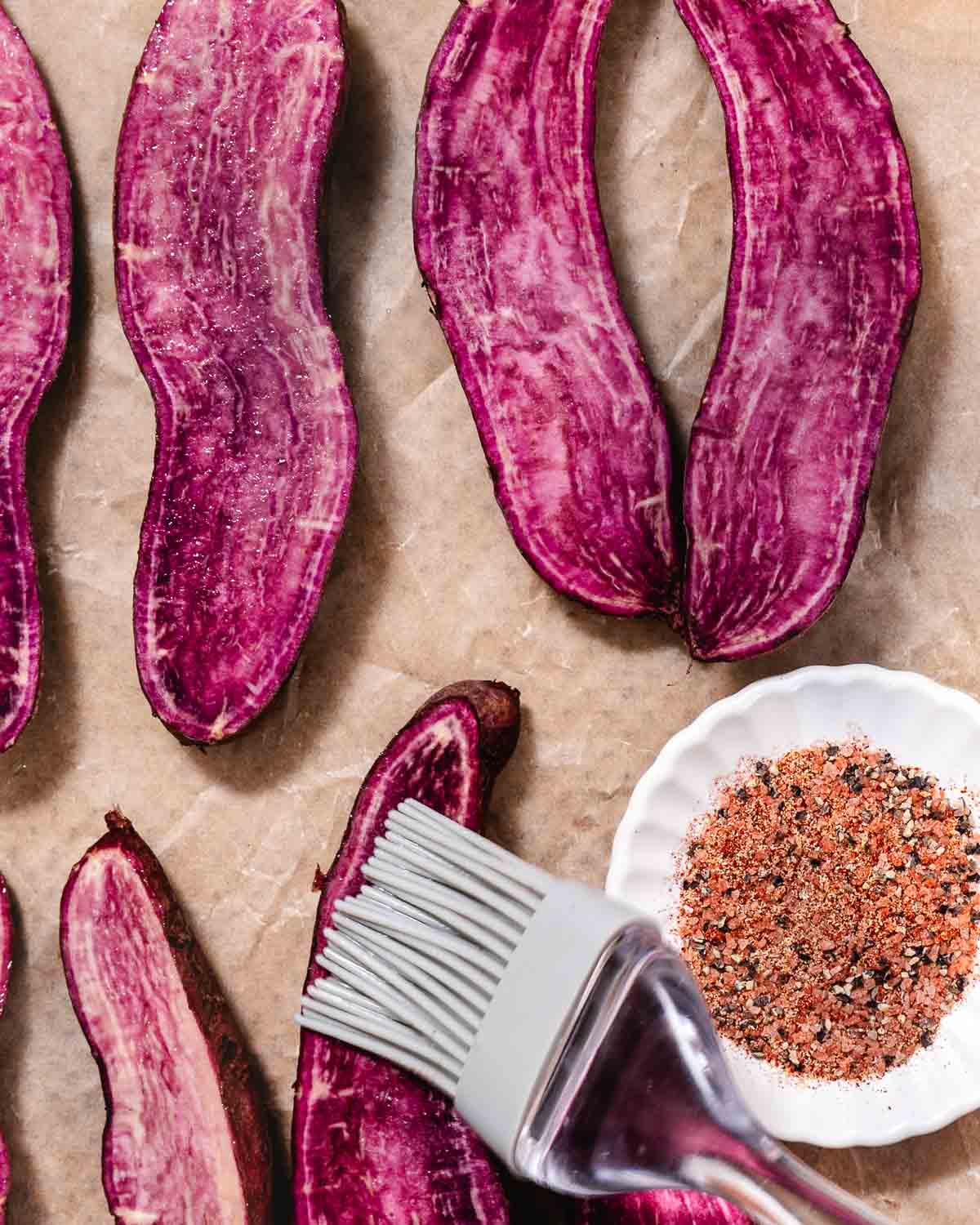 Brushing oil onto halved purple sweet potatoes before seasoning and roasting.