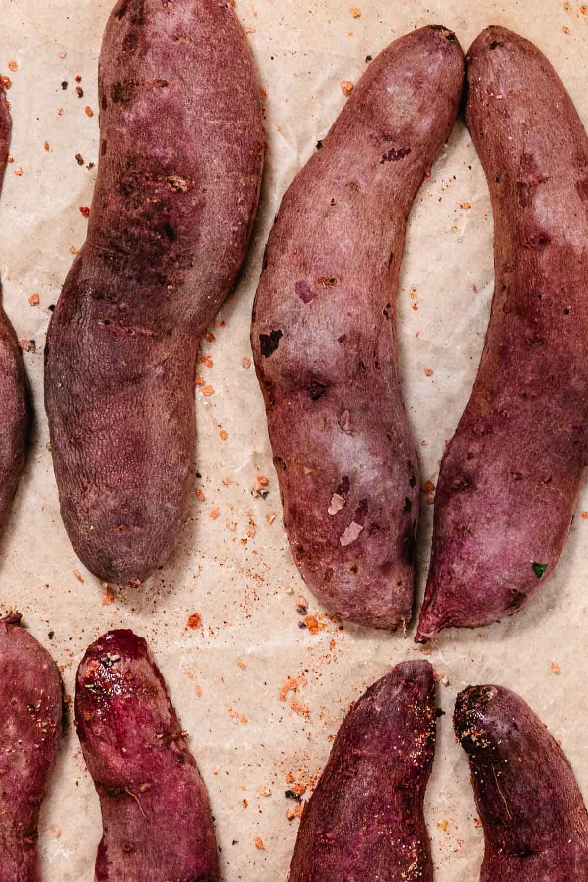 Whole purple sweet potatoes and halved purple sweet potatoes on a parchment-lined baking sheet before roasting.