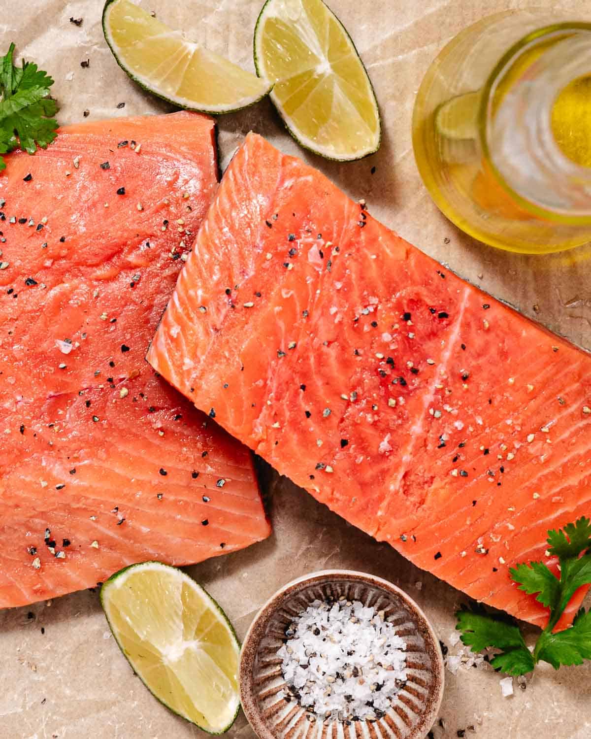 Raw coho salmon fillets seasoned with salt and black pepper, surrounded by lime wedges, olive oil, and fresh cilantro before cooking.