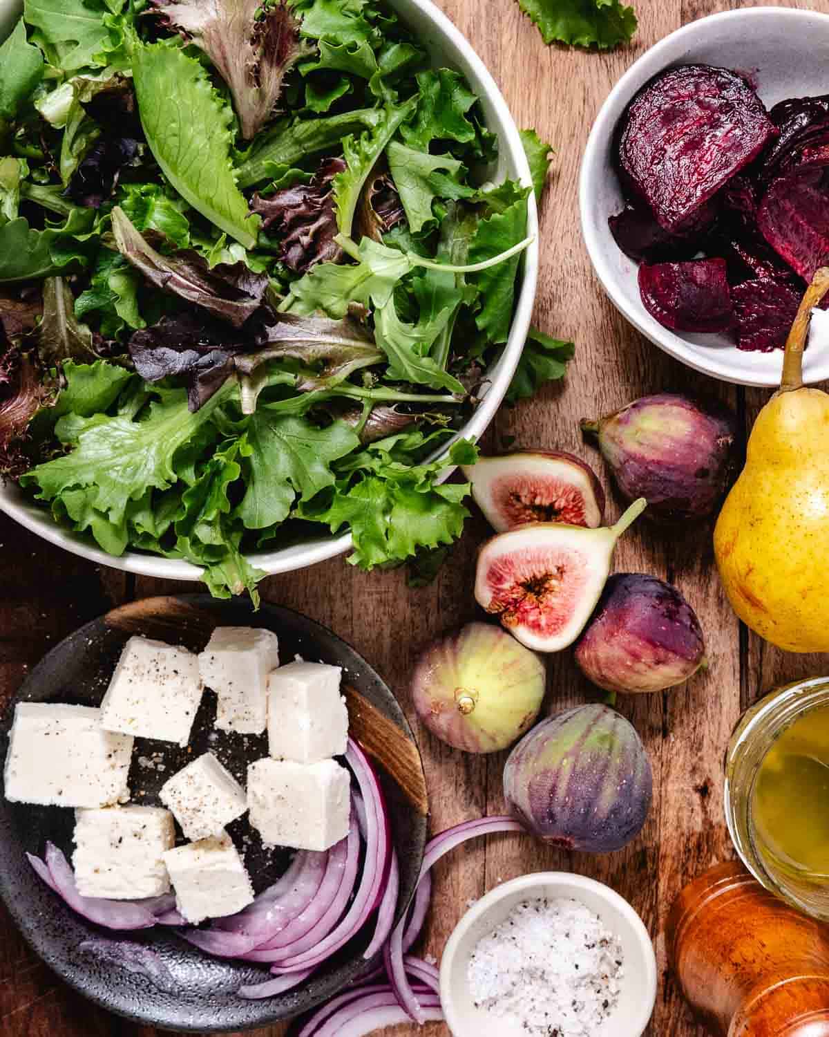 Ingredients for beet and pear salad arranged on a wooden table, including mixed greens, roasted beets, pears, figs, feta, almonds, red onion, and lemon Dijon dressing.