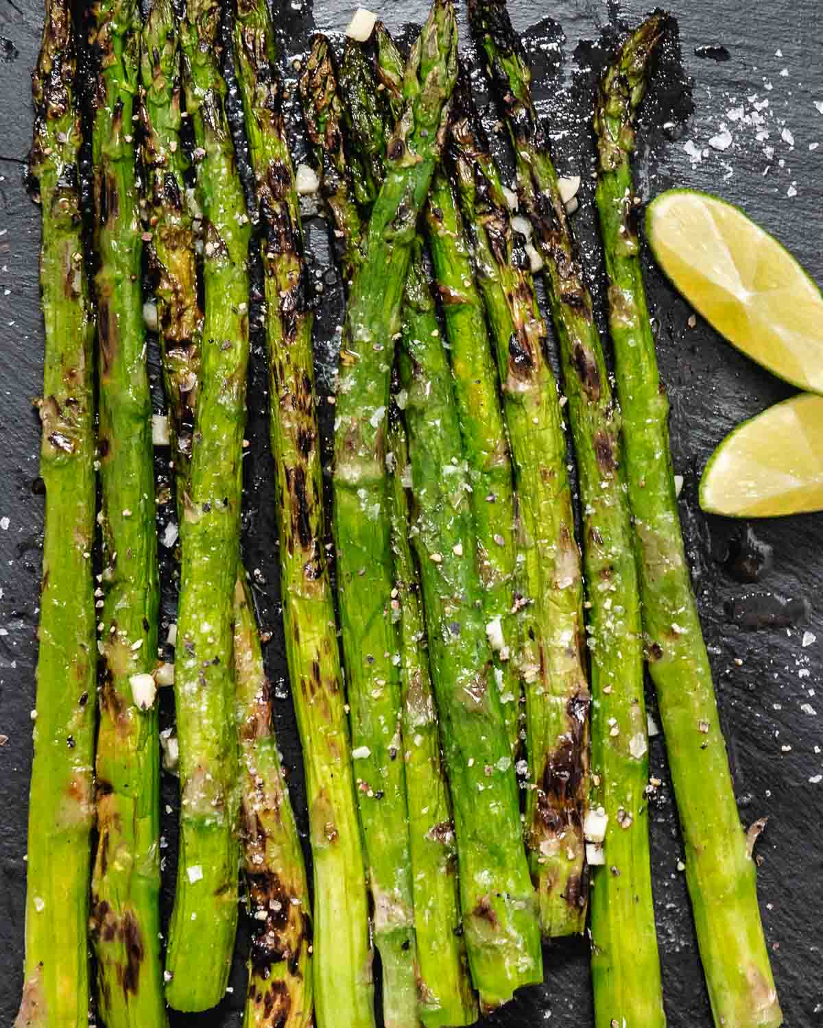 Grilled asparagus with citrus wedges on a black plate.