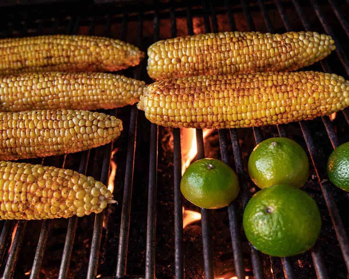 Grilled corn on the cob seasoned with Old Bay cooking over an open grill with fresh limes.