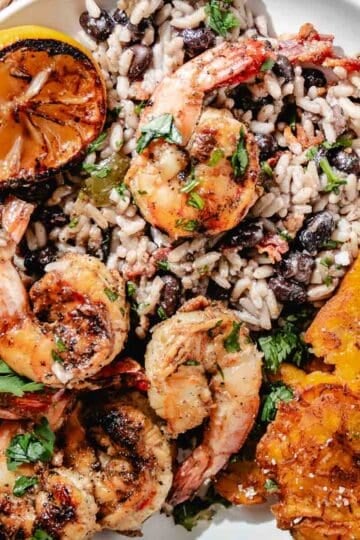 Grilled shrimp with rice and black beans, fried plantains, cilantro, and charred citrus on a white plate.