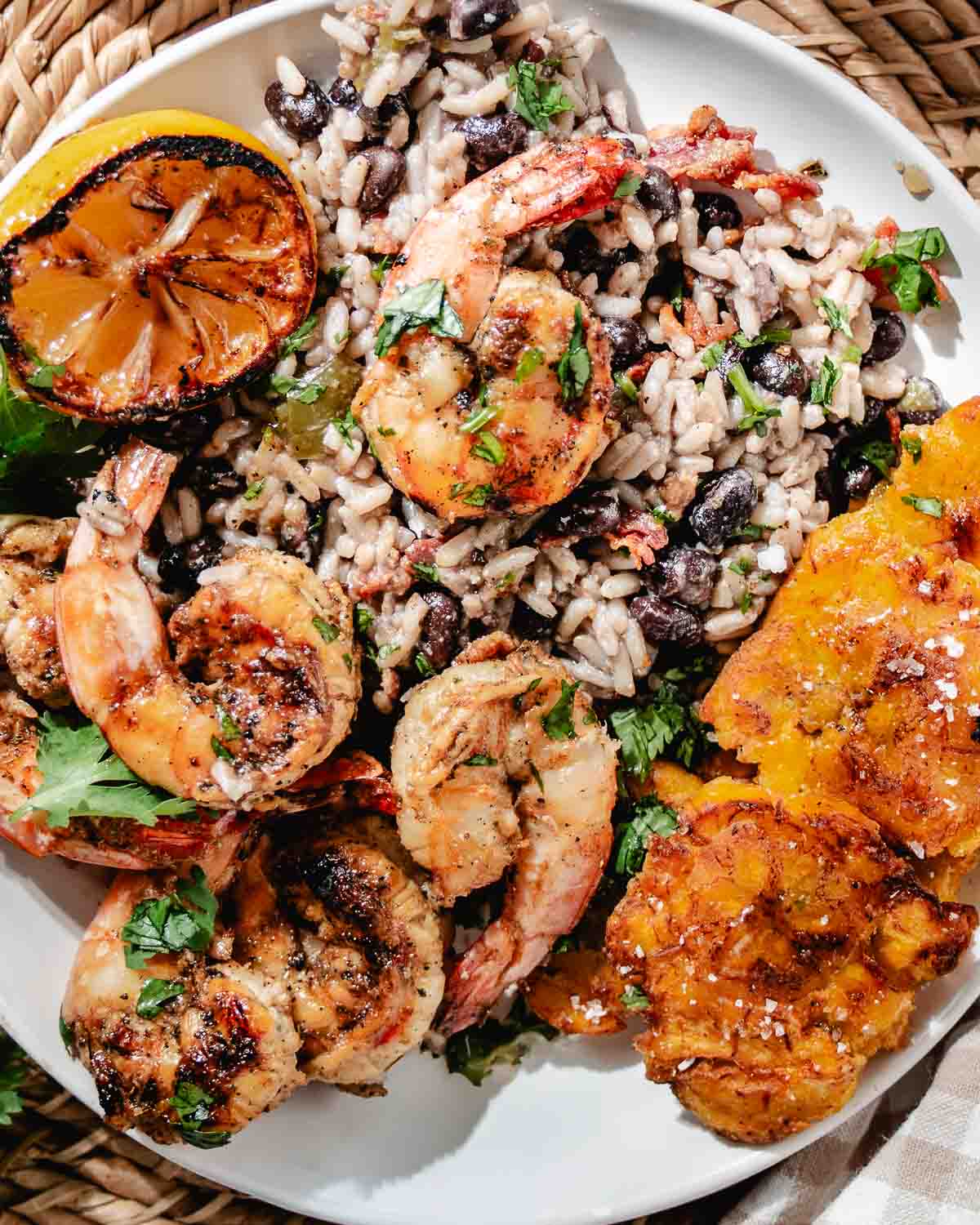 Grilled shrimp with rice and black beans, fried plantains, cilantro, and charred lemon on a white plate.