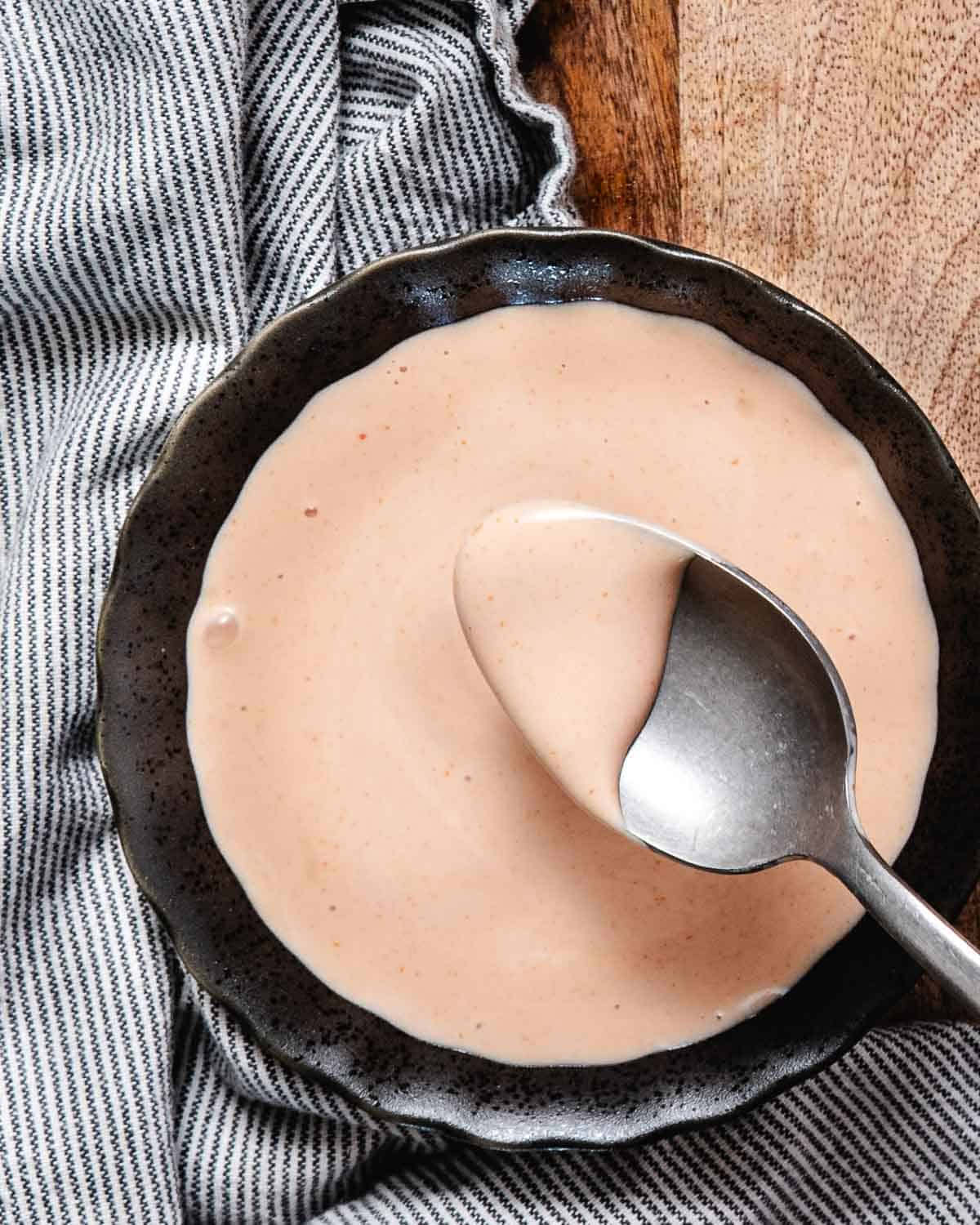 Spoon lifting creamy Marie Rose sauce from a dark bowl on a striped kitchen towel.