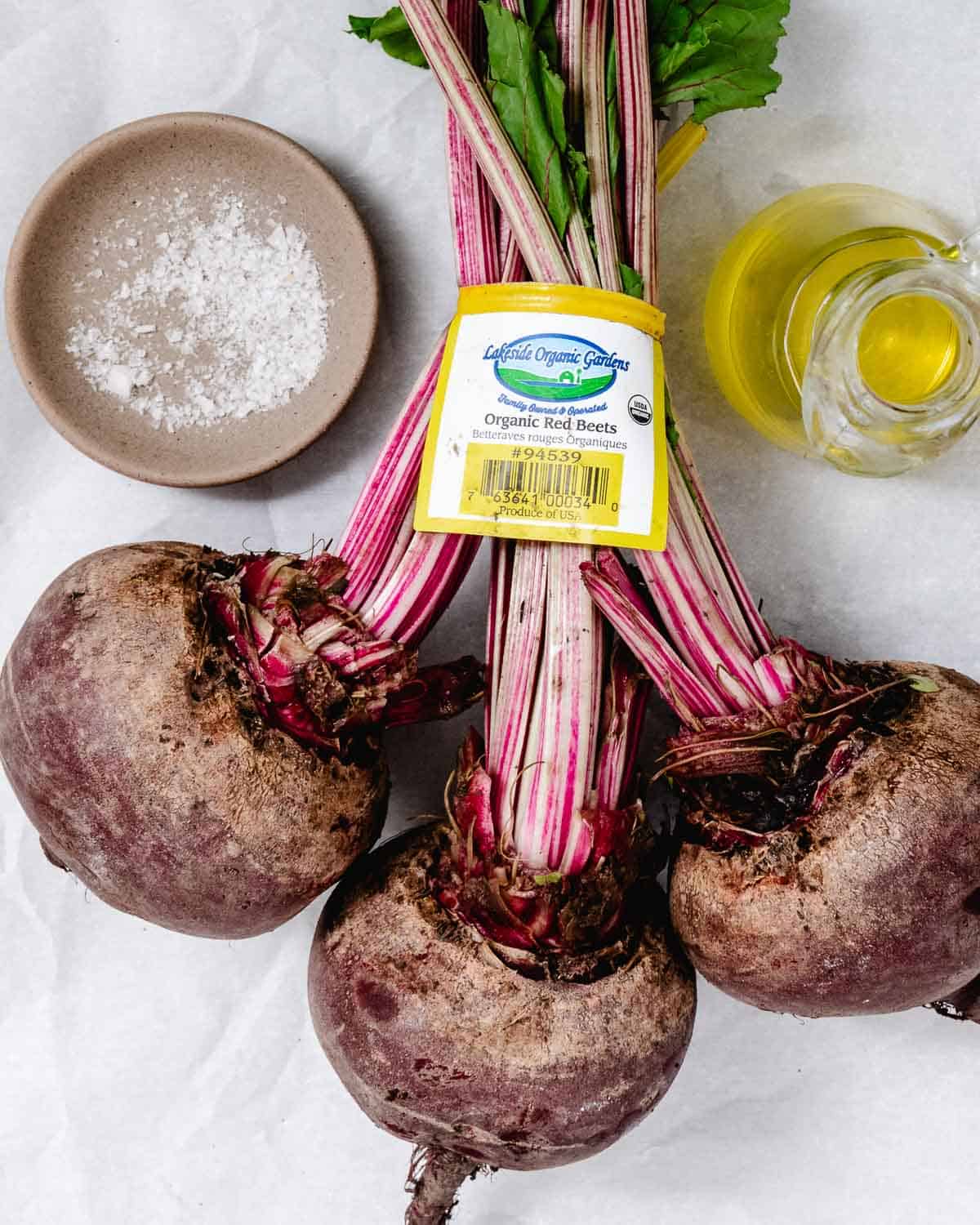 Fresh red beets with leafy stems, olive oil, and kosher salt on a white surface.