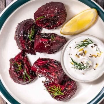 Roasted red beets on a white plate with dill yogurt sauce and a lemon wedge.