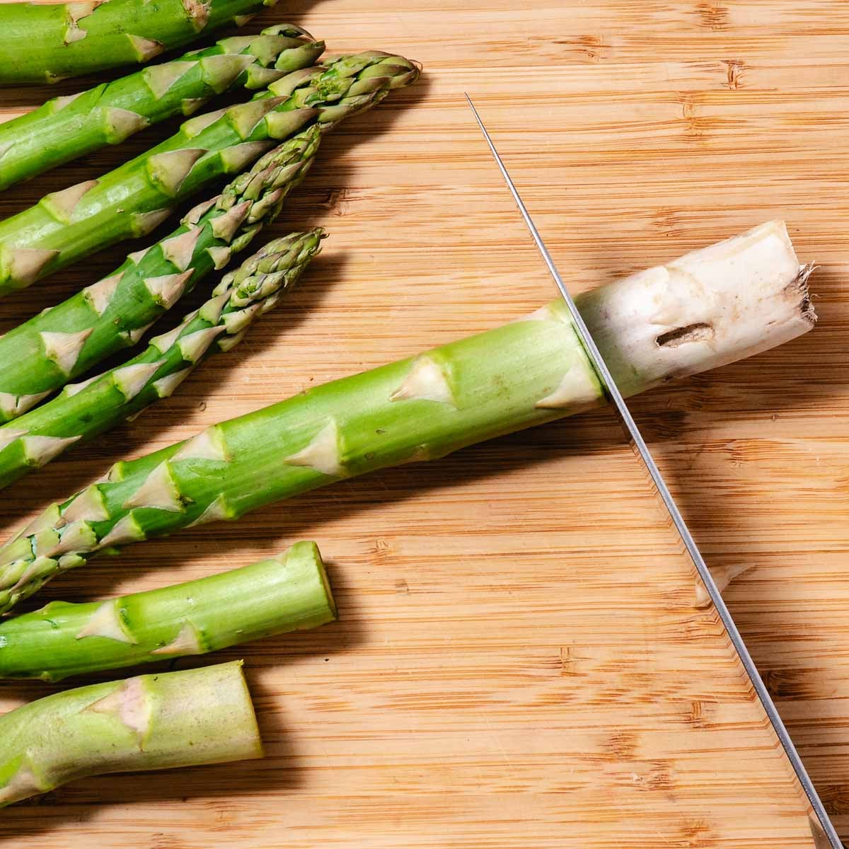 Thick asparagus spear being peeled flat on a wooden cutting board with a swivel vegetable peeler.