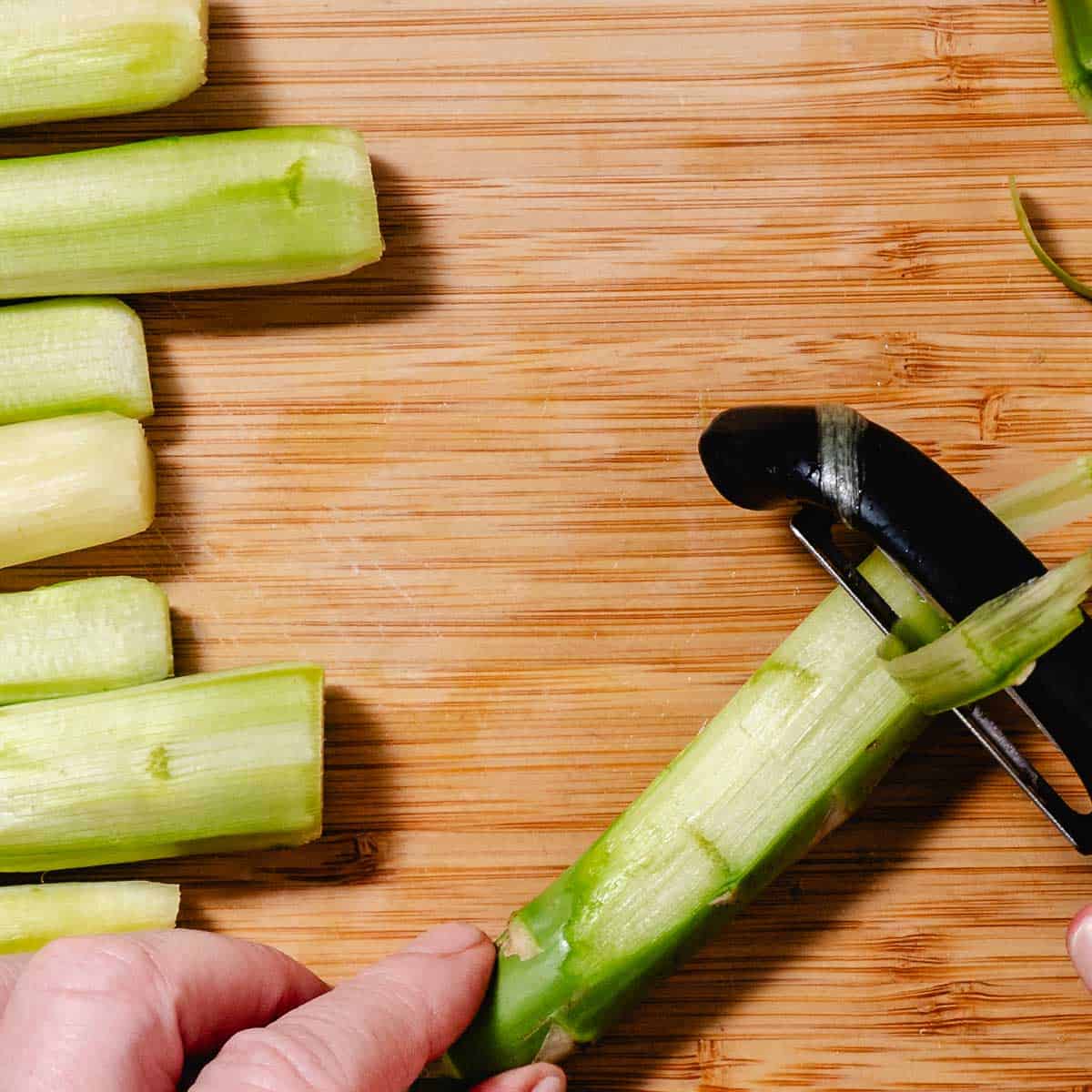 Thick asparagus spear being peeled flat on a wooden cutting board with a swivel vegetable peeler.