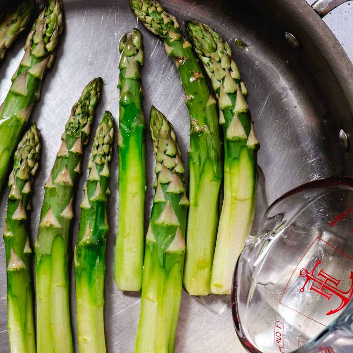 Thick asparagus spears cooking with water in a covered skillet.