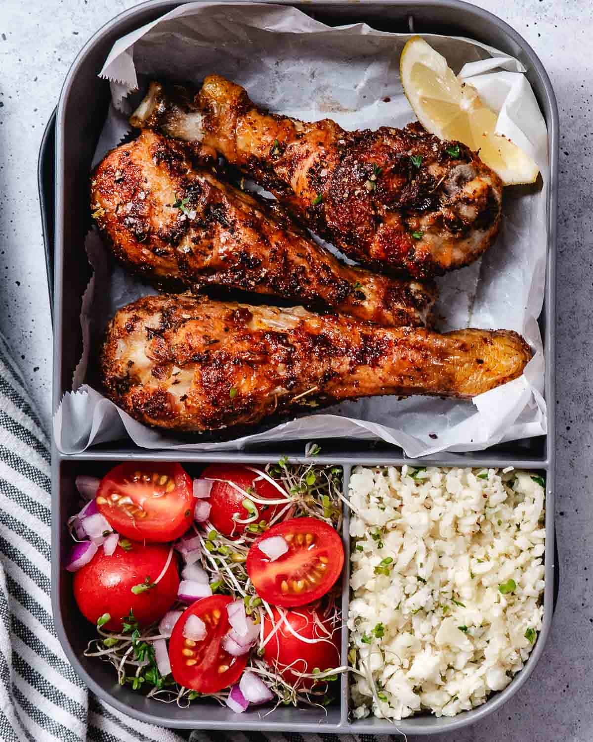 Air fryer chicken drumsticks with cauliflower rice and tomato salad in a meal-prep container.