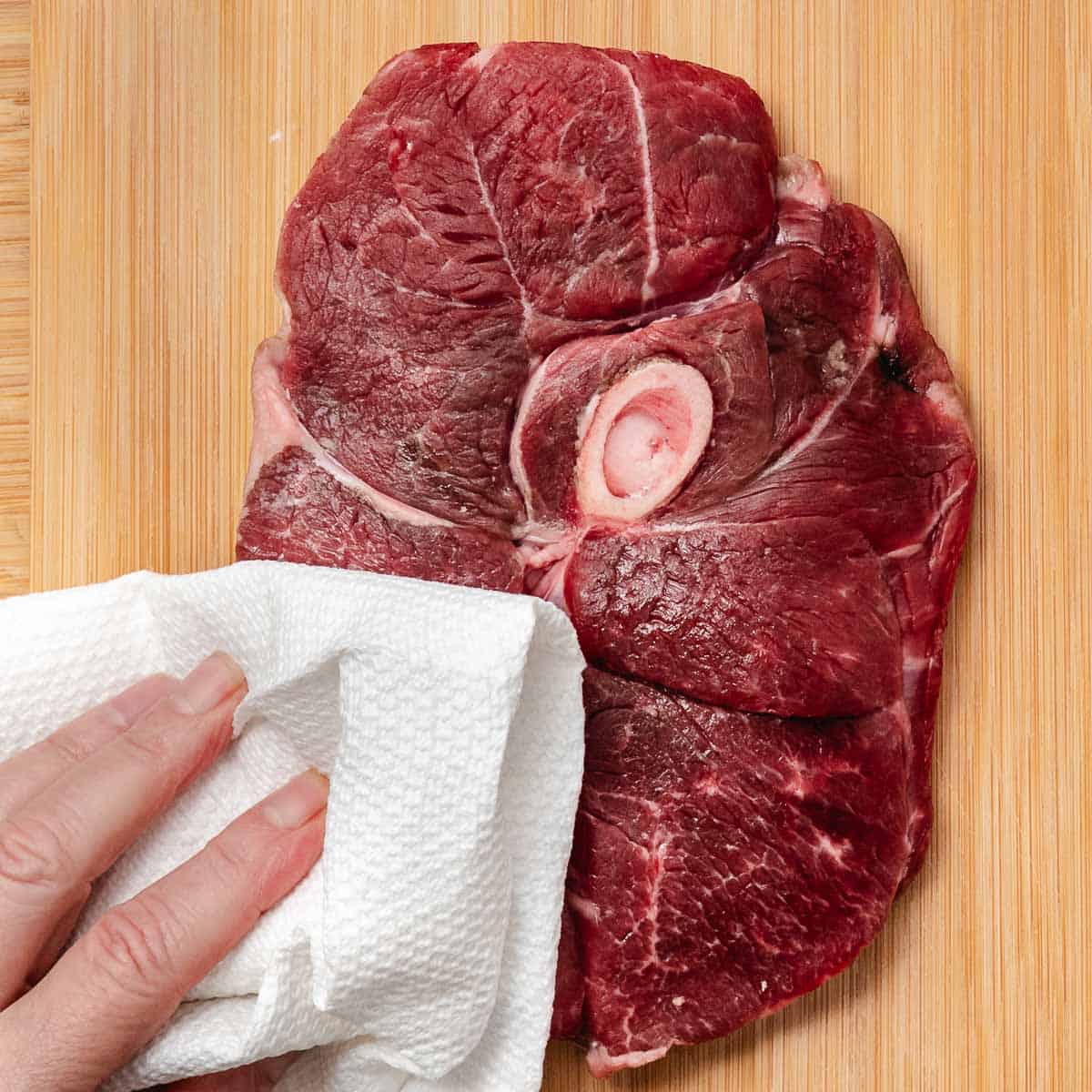 Patting a raw bone-in lamb leg steak dry with a paper towel on a wooden cutting board.