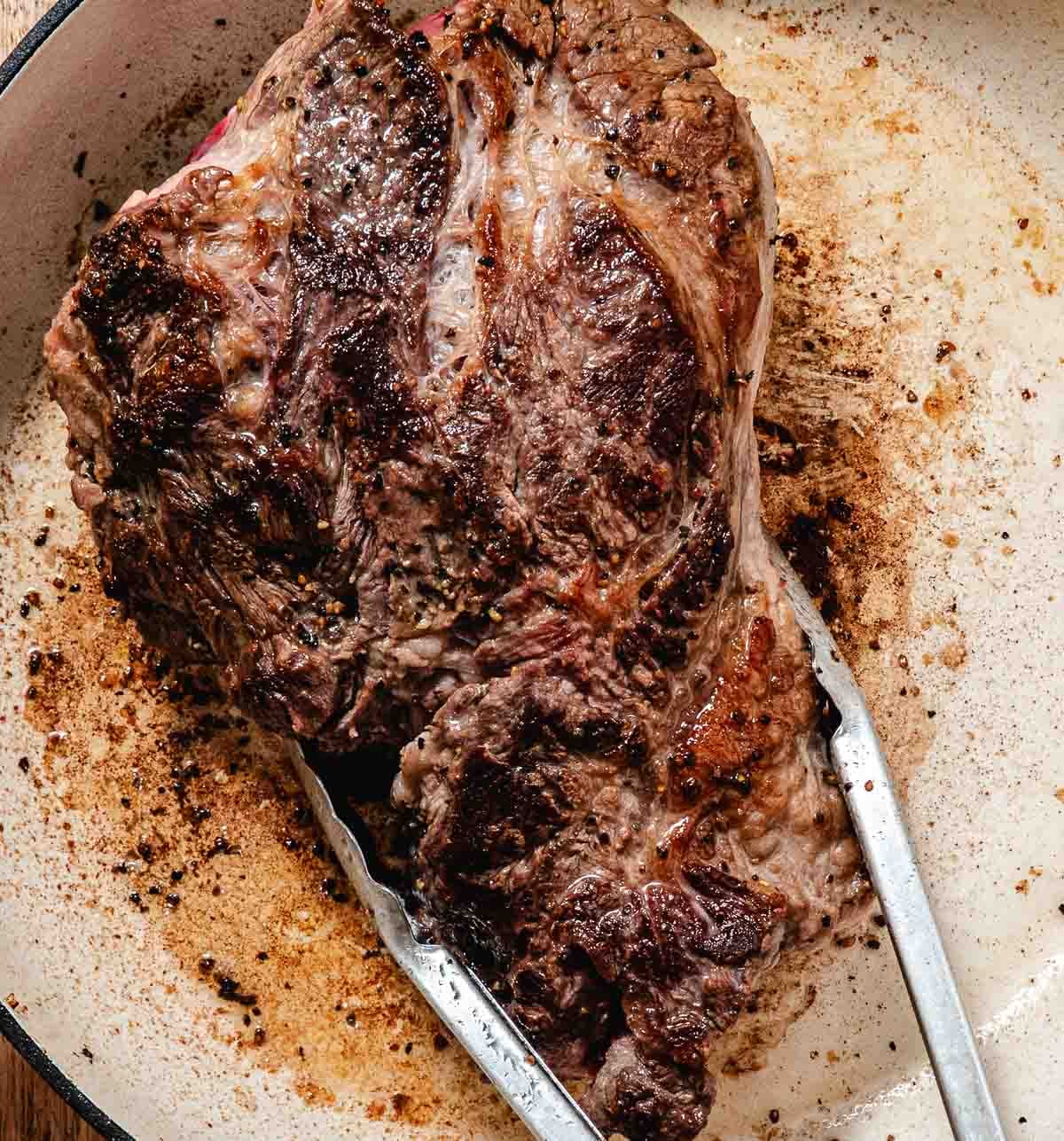 Chuck roast browning in a skillet with metal tongs.