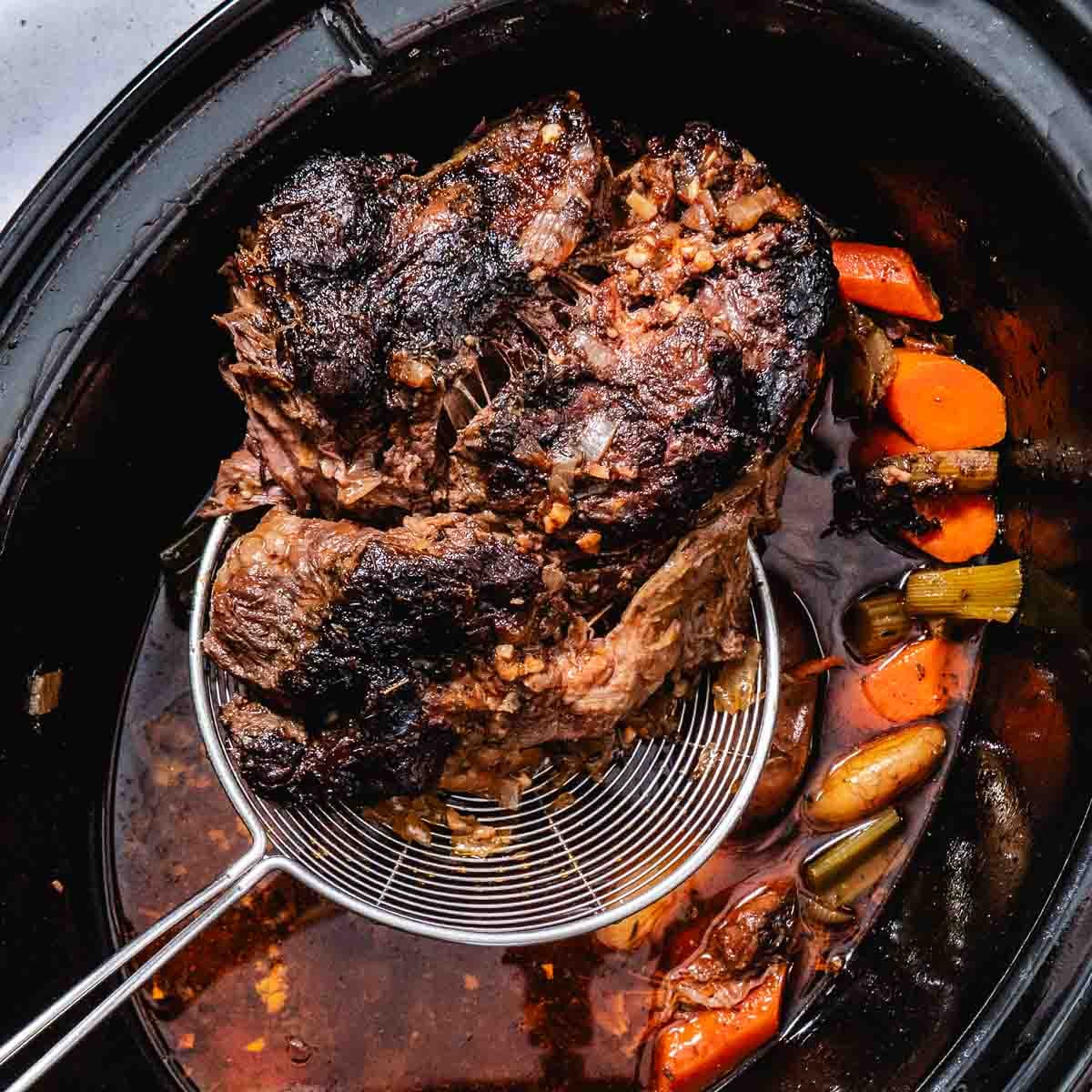 Cooked chuck roast being lifted from a slow cooker with a wire skimmer.