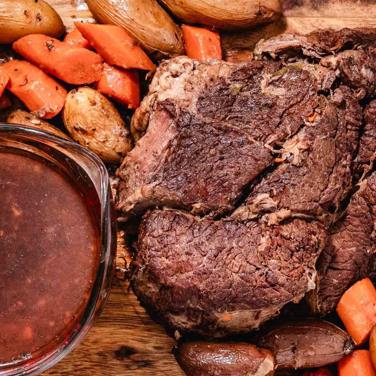 Cooked chuck roast and vegetables beside a glass container of strained cooking liquid.