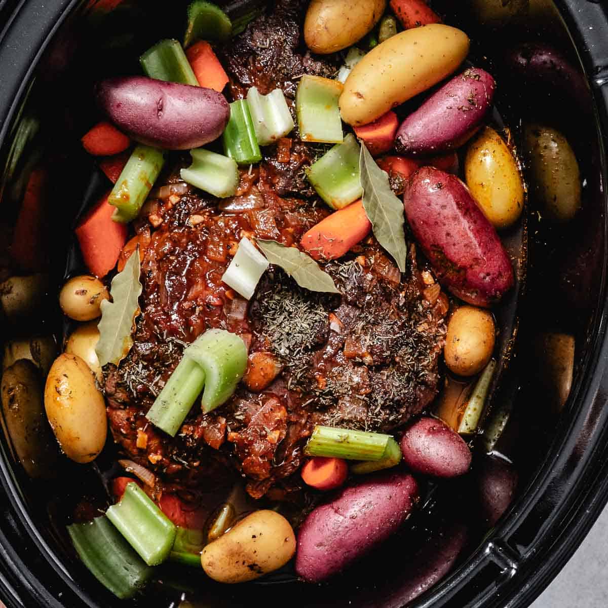 Browned chuck roast with potatoes, carrots, celery, bay leaves, and thyme in a slow cooker before cooking.