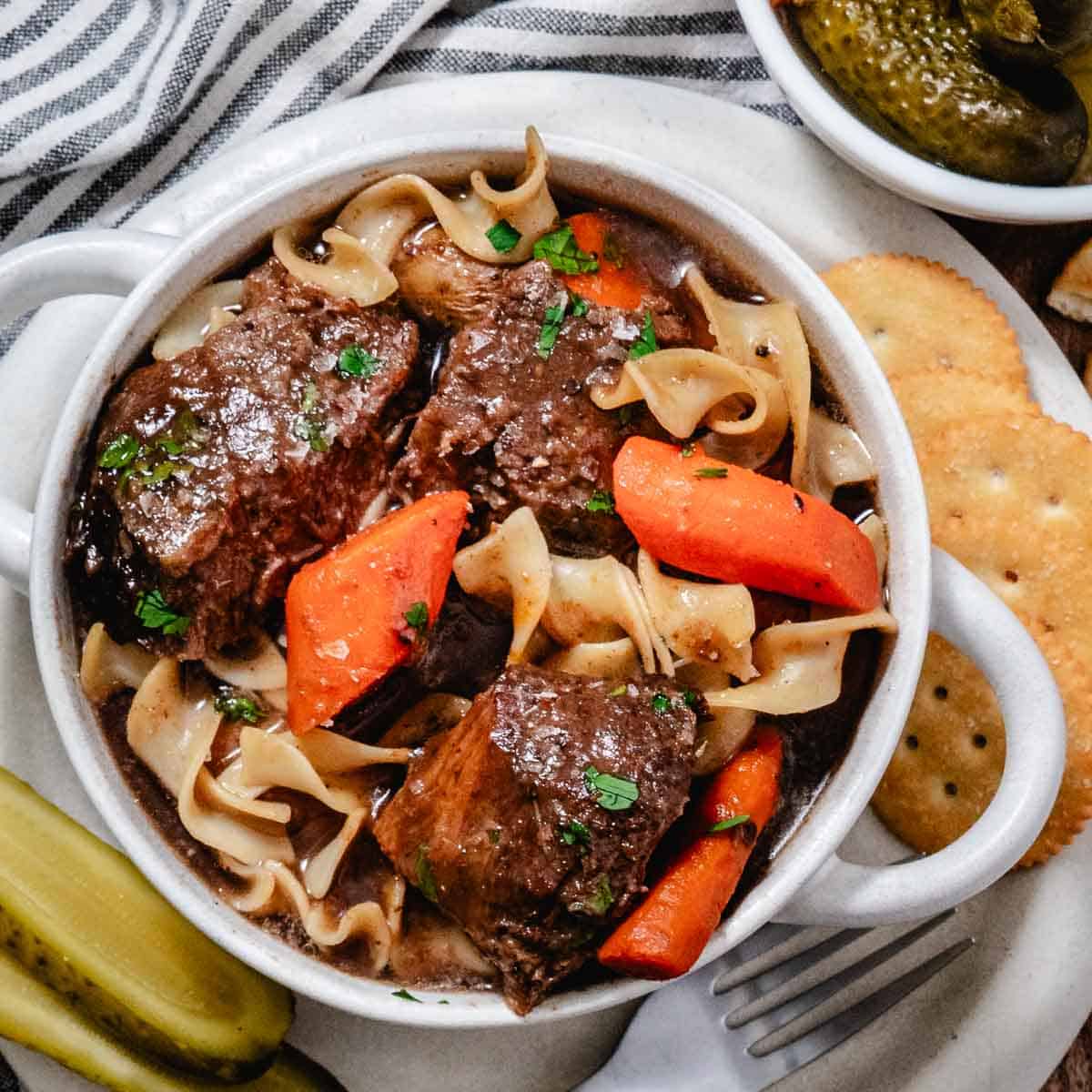 Chuck roast with carrots, egg noodles, and gravy in a white bowl, served with crackers and pickles.