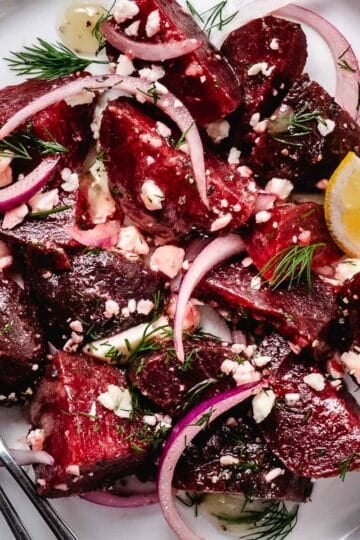 Beet salad with feta cheese, red onion, and fresh dill on a white plate with a lemon wedge and fork.