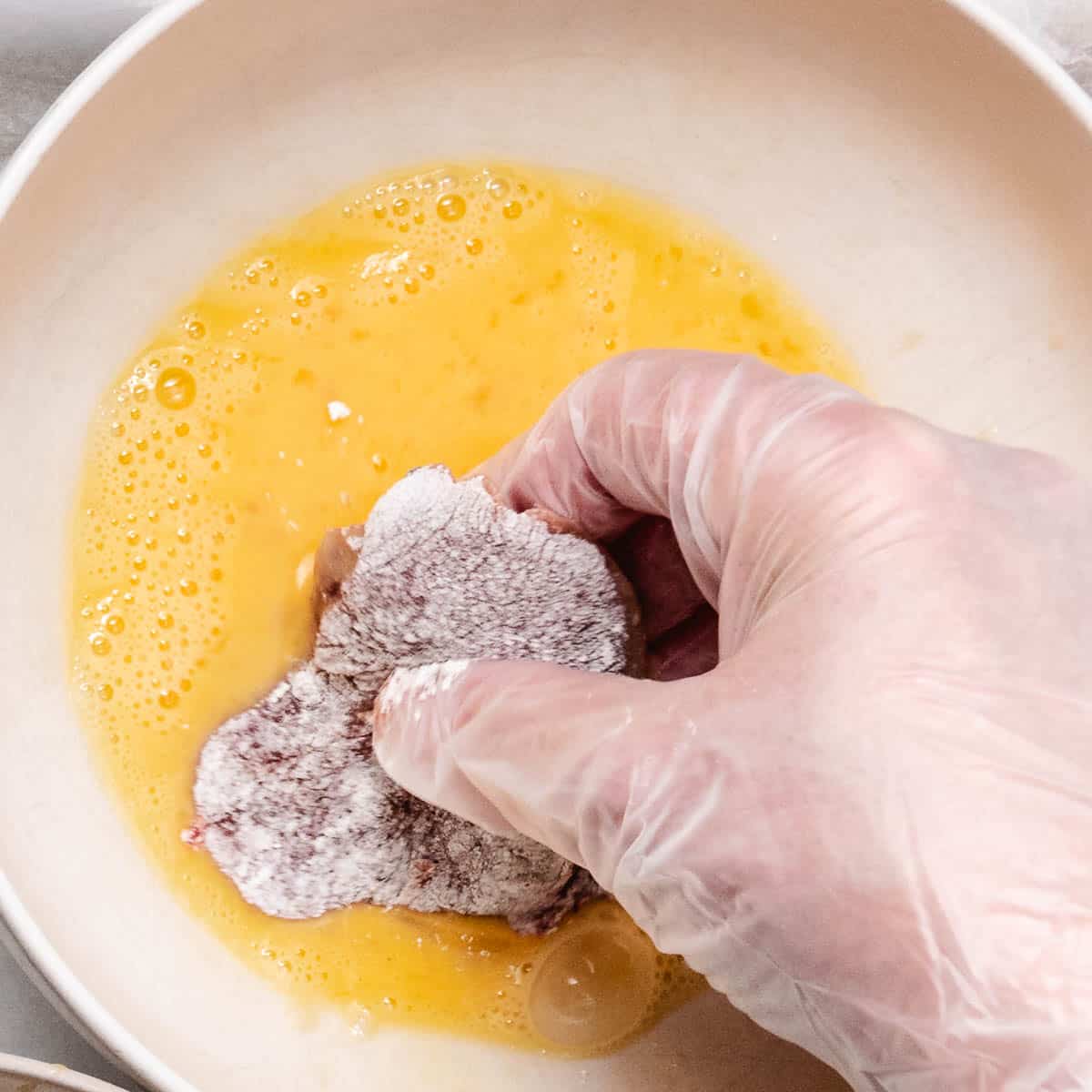 A gloved hand dipping a flour-coated chicken liver into beaten eggs.