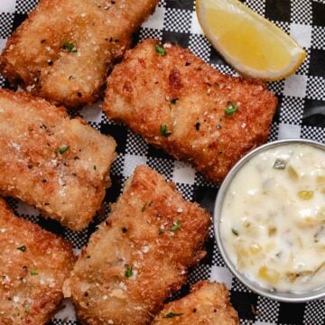Crispy pan-fried pollock fillets on black-and-white checkered paper with lemon and tartar sauce.