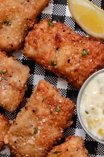 Crispy pan-fried pollock fillets on black-and-white checkered paper with lemon and tartar sauce.